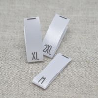 N&auml;rbild p&aring; tre vita v&auml;vda storleksetiketter i satin med tryckta storlekar M, XL och 2XL, fr&aring;n artikeln Storlekstag XL &ndash; V&auml;vd Etikett i Satin.