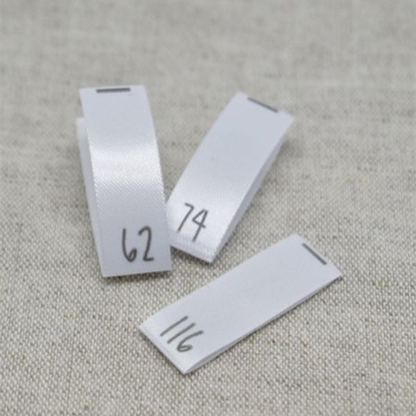 N&auml;rbild p&aring; tre vita v&auml;vda storleksetiketter i satin med tryckta siffror 62, 74 och 116, fr&aring;n artikeln Storlekstag 056 &ndash; V&auml;vd Etikett i Satin.