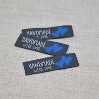 N&auml;rbild p&aring; tre svarta v&auml;vda etiketter med vit text "Handmade with love" och tv&aring; bl&aring; hj&auml;rtan, fr&aring;n artikeln V&auml;vd Etikett Handmade With Love Bl&aring; &ndash; 10-pack.